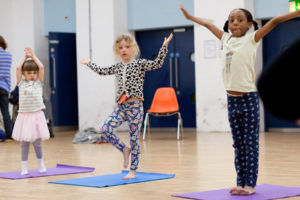 Three young girls are standing on yoga mats and holding their arms up in the air.