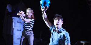 One Small Step Production photo. In the foreground a man with short dark hair wearing a blue shit and black bow tie is holding up a black and blue football. Behind him a woman in a black and white striped tshirt is holding a toy spaceship.