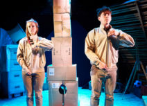 One Small Step production image. A man and a woman dressed in beige are facing forward, zipping up their jackets in unison. In the background is a tower made of cardboard boxes.