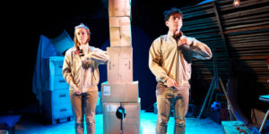 One Small Step production image. A man and a woman dressed in beige are facing forward, zipping up their jackets in unison. In the background is a tower made of cardboard boxes.