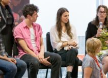 Picture of five adults at Z-arts. Four are sitting on chairs and one is sitting in front of them on the floor looking behind at them. One of the adults on the chairs is speaking into a microphone.