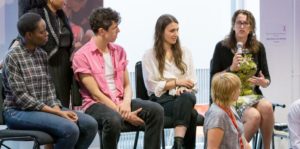 Picture of five adults at Z-arts. Four are sitting on chairs and one is sitting in front of them on the floor looking behind at them. One of the adults on the chairs is speaking into a microphone.