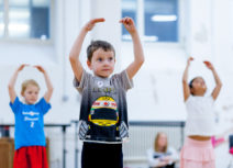 Photo of young ballet dancers. A boy wearing a grey tshirt with a racing driver on the front is holding his arms above his head. Two other girls are doing the same action in the background.
