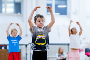 Photo of young ballet dancers. A boy wearing a grey tshirt with a racing driver on the front is holding his arms above his head. Two other girls are doing the same action in the background.