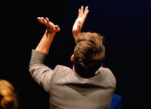 A man with short brown hair wearing a grey blazer is facing away from the camera and clapping into the air.