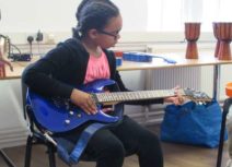 A girl is sat wearing a black cardigan, pink tshirt and black leggings playing a blue electric guitar.