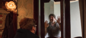 Production photo from INUK. A man is pressing his hands and face up against a glass panel on a door.