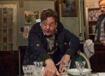 Production image from INUK. An older man is frantically setting a table in the foreground, behind him, a younger man wearing a navy coat and a yellow beanie hat is watching him with a puzzled expression on his face.