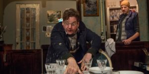 Production image from INUK. An older man is frantically setting a table in the foreground, behind him, a younger man wearing a navy coat and a yellow beanie hat is watching him with a puzzled expression on his face.