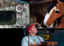 Production photo from Black Beauty. A man holding a bucket wearing a red headband and blue tshirt is looking up at a brown and white pantomime horse who is inside a blue horsebox.