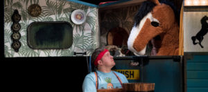 Production photo from Black Beauty. A man holding a bucket wearing a red headband and blue tshirt is looking up at a brown and white pantomime horse who is inside a blue horsebox.
