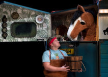 Production photo from Black Beauty. A man holding a bucket wearing a red headband and blue tshirt is looking up at a brown and white pantomime horse who is inside a blue horsebox.