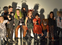 Children and an artist stand in a line in different halloween costumes making scary faces at the camera