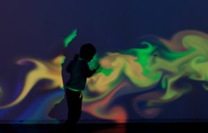 Photo of a colourful, psychelic style projection of yellow, green, red and blue on a wall silhouetting a young child standing in front of it.