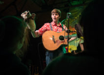 Photo of a young man with short brown hair wearing a red and white patterned shirt holding a puppet and a guitar