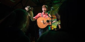 Photo of a young man with short brown hair wearing a red and white patterned shirt holding a puppet and a guitar
