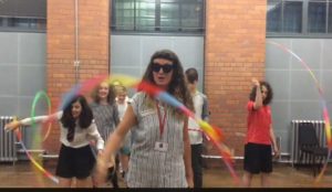 A woman wearing sunglasses and a stripey, sleeveless shirt is waving a colourful ribbon. Behind her, young teenagers are playing with circus equipment.