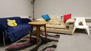 Photo of a room with two sofas, one blue and one cream. There is a white plastic chair next to the cream sofa and a black, grey and pink rug between the two sofas. On top of the rug is a small round coffee table.