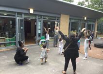 Parents and children dancing together outside. They are all holding their hands up in front of the air and are standing in front of a school building in a playground