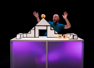 Production photo of A Square World. Daryl Beeton with an angular set on a table, with two squares and one circle. The circle is stuck at the top of the angular set.