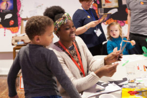 Z-arts Engagement Officer Yemi is wearing a cream cardigan, a red lanyard and a colourful head band. She is sitting at a table and smiling at a young boy wearing a grey long-sleeved shirt who is standing next to her.
