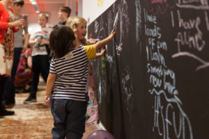 Two children are standing in front of a wall which has been painted with chalkboard paint. They are both drawing on the wall. One child is wearing long blonde hair and a pink, floral dress. The other child has short dark, brown hair and a blue and white-striped t-shirt.