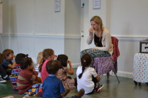 A woman with blonde hair is sitting on a chair holding her nose. A group of children are sitting in front of her and looking at her, copying her by holding their own noses.