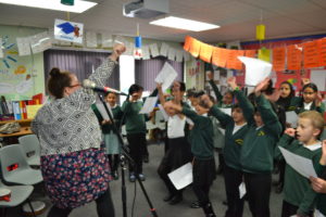 Pupils and artist taking part in a school workshop. An artist is standing in front of a group of children wearing green school jumpers, she is standing next to a microphone and punching the air, the children are copying her movements.