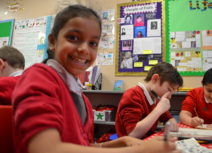 A young girl wearing a red school cardigan is looking directly at the camera and smiling, two of her schoolmates wearing red jumpers are in the background drawing on pieces of paper