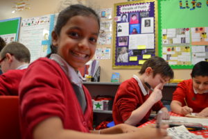 A young girl wearing a red school cardigan is looking directly at the camera and smiling, two of her schoolmates wearing red jumpers are in the background drawing on pieces of paper