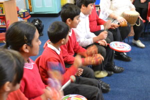 Children wearing red school jumpers are sitting and playing musical instruments including, drums, maracas and bongo drums