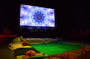 Z-arts sensory space, a psychedelic pattern is projected onto the wall of a dark space and the floor is illuminated in green. There are pillows and different textured fabrics on the floor.