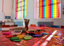 Lead image for About Us page. Photo of Z-arts gallery workspace. A table is covered in paint-stained shiny red tablecloths with cardboard, felt, paintbrushes and other art resources on it. In the background there are rainbow coloured blinds covering two windows.