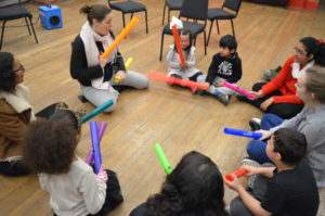 A group of children and adults are sat cross-legged on the floor in a circle, they are all holding different brightly-coloured plastic tubes.