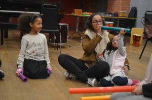 A woman wearing a brown coat is blowing into a turquoise tube, she has her arm wrapped round her daughter who is wearing a pink striped gilet and grey polkadot jumper. A girl wearing a grey jumper and leopard-print cat ears looks on.