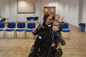 Photo of young girl with blonde hair wearing planet-patterned leggings sitting on the arm-rest of a brown-haired woman's wheelchair.