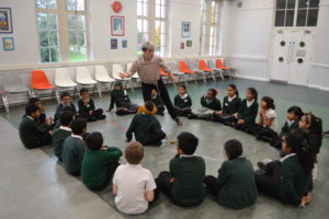 An adult with short grey hair and black trousers is standing in the centre of a group of school children leading a physical workshop.