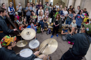 A man playing a clarinet, a man playing on a drum kit and a man playing a double bass perform in front of a group of children and families