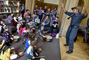 A man wearing a blue jumpsuit and purple and blue-striped tie is dancing and holding a clarinet between his head and shouler. In front of him families and children are sat playing on musical instruments including drums and rainbow coloured xylophones.