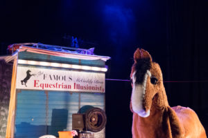 A photo of Hamish the pantomime horse from Black Beauty. The horse is brown with a white nose and long eyelashes and he is standing in front of a horsebox which reads 'The Famous McCuddy brother's Equestrian Illusionists'
