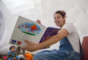 A woman with brown hair wearing denim dungarees and a white t-shirt is sitting reading a book to a group of children.