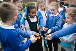 Several schoolchildren are standing in a circle, they are all holding coloured pencil crayons and putting their hands into the centre of the circle