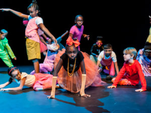 Photo of children crawling on the floor, at the centre of the image is a girl looking directly at the camera wearing an orange bow, necklace, long gloves, net skirt and a black top.
