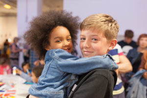 A boy is holding a little girl and she has her arms wrapped round his neck, they are both looking directly at the camera.