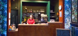 Lead picture for Job Opportunities page - A staff member wearing a red Z-arts polo shirt stands behind the wooden counter at Z-cafe, In the background is a coffee machine, jars with different types of teas, wine and pint glasses, a selection of fruit and various other food items.