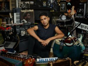A man is sat on the floor surrounded by different musical instruments including drums, keyboards, accordians, guitars and musical speakers, headphones and amps.