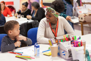 Z-arts Engagement Officer Yemi is wearing a cream cardigan and a colourful head band. She is sitting at a table and smiling at a young boy wearing a grey long-sleeved shirt sitting next to her. There are felt tip pens and scissors on the table.