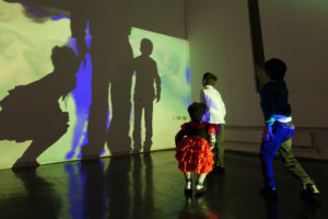 Children dancing and watching their shadows projected onto a wall by yellow, blue and green light.