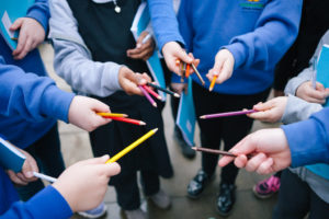 Several schoolchildren are standing in a circle, they are all holding coloured pencil crayons and putting their hands into the centre of the circle
