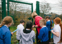 A teacher and a group of children are looking closely at a patch of grass behind a green metal mesh fence. The children are all holding light blue workbooks.